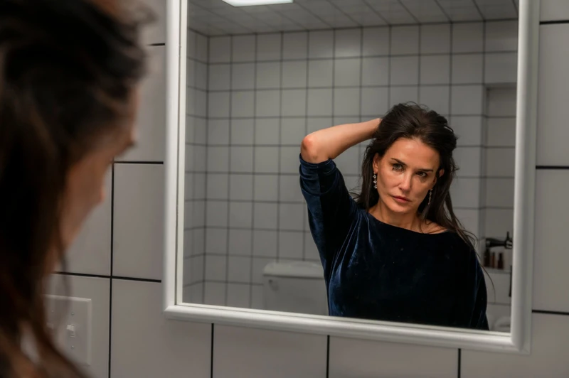 Personagem de Demi Moore em A Substância olhando-se no espelho com roupas escuras.