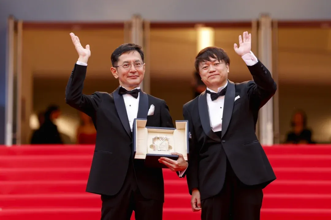 Dois homens de terno em um tapete vermelho segurando um prêmio com duas mãos acenando.