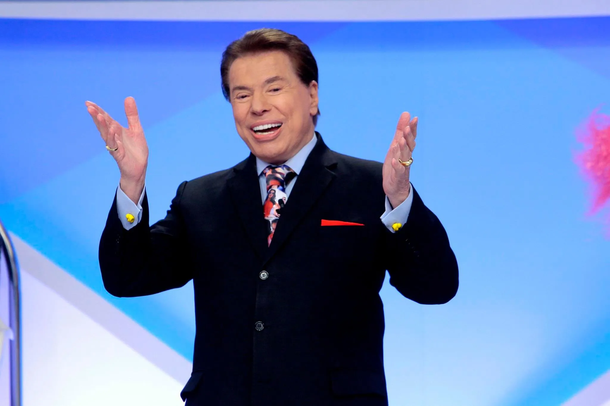 Silvio Santos, em um fundo azul, sorridente, utilizando terno e um microfone acoplado.