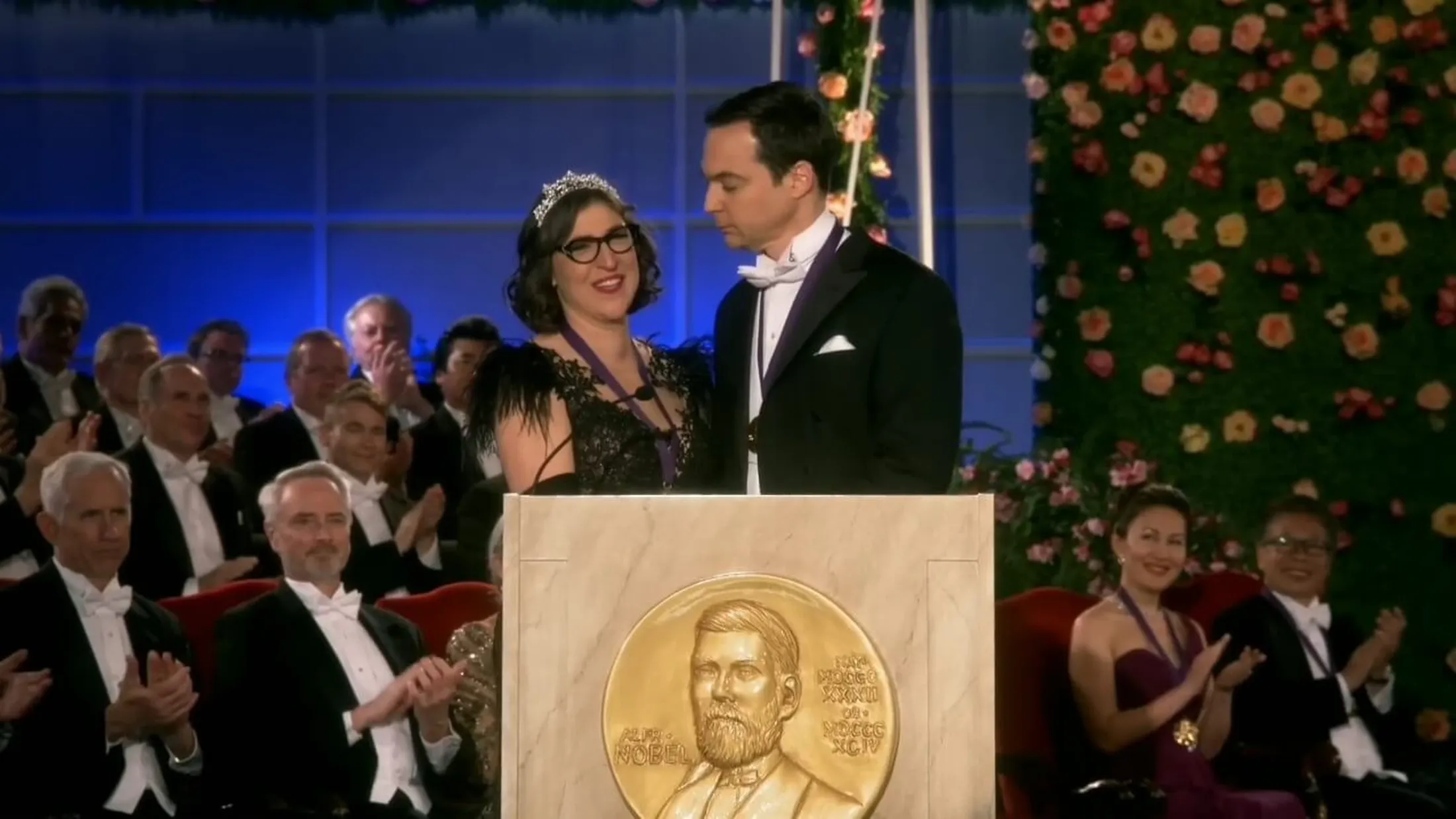 Amy de tiara usando vestido preto e Sheldon de terno na premiação do Nobel