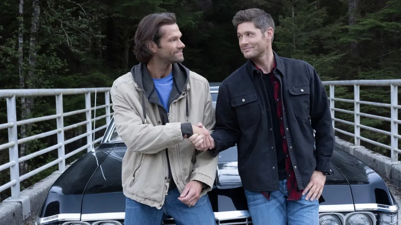 Os irmãos Winchester, da mesma altura, em pé um do lado do outro sorridente e com as mãos dadas em um aperto.