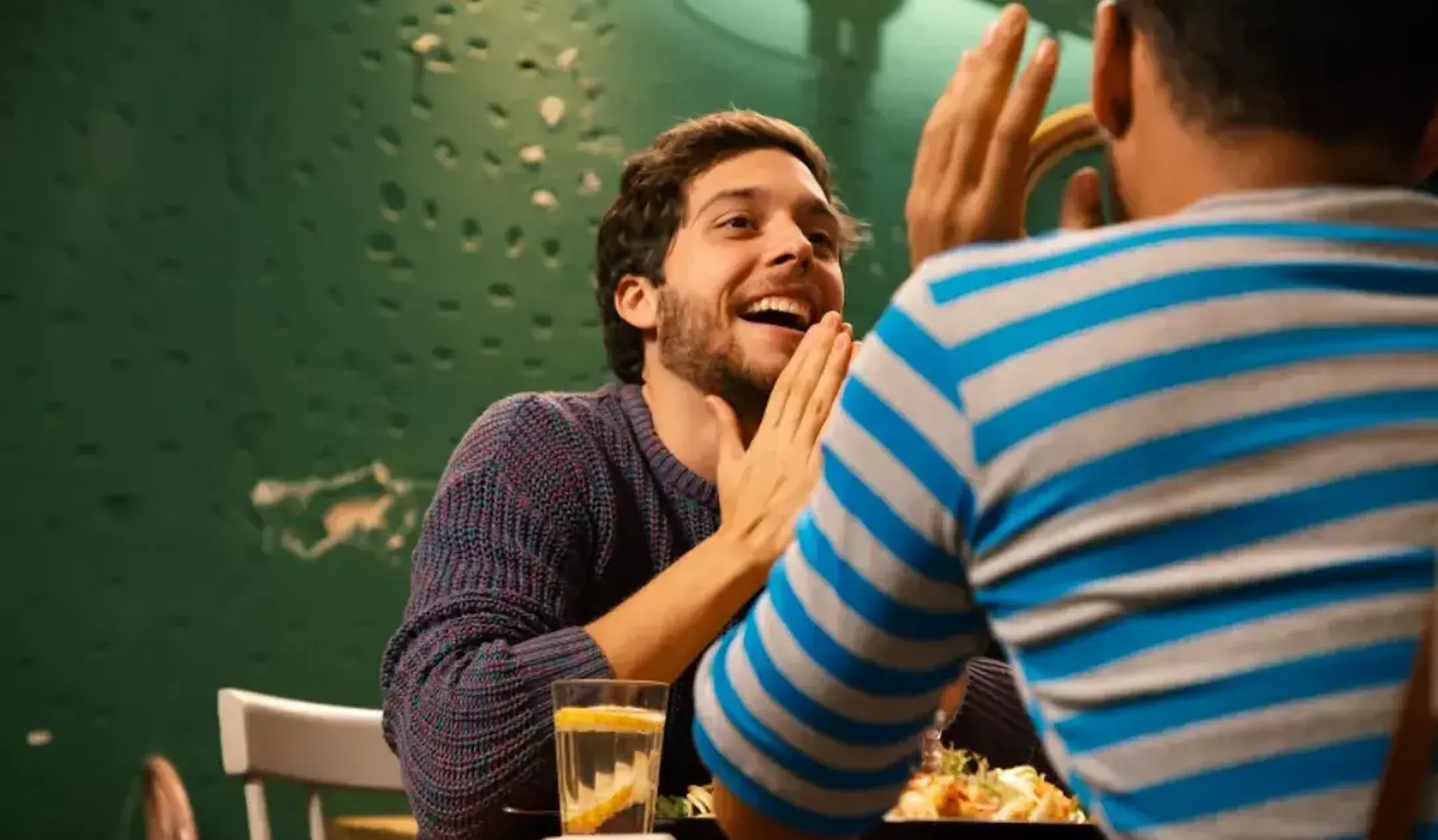 Protagonista de 13 Sentimentos sorridente de frente para outra pessoa, sentado à uma mesa.