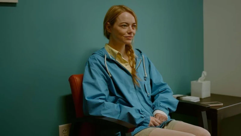 Emma Stone em Tipos de Gentileza sentada com jaqueta azul e sorridente.