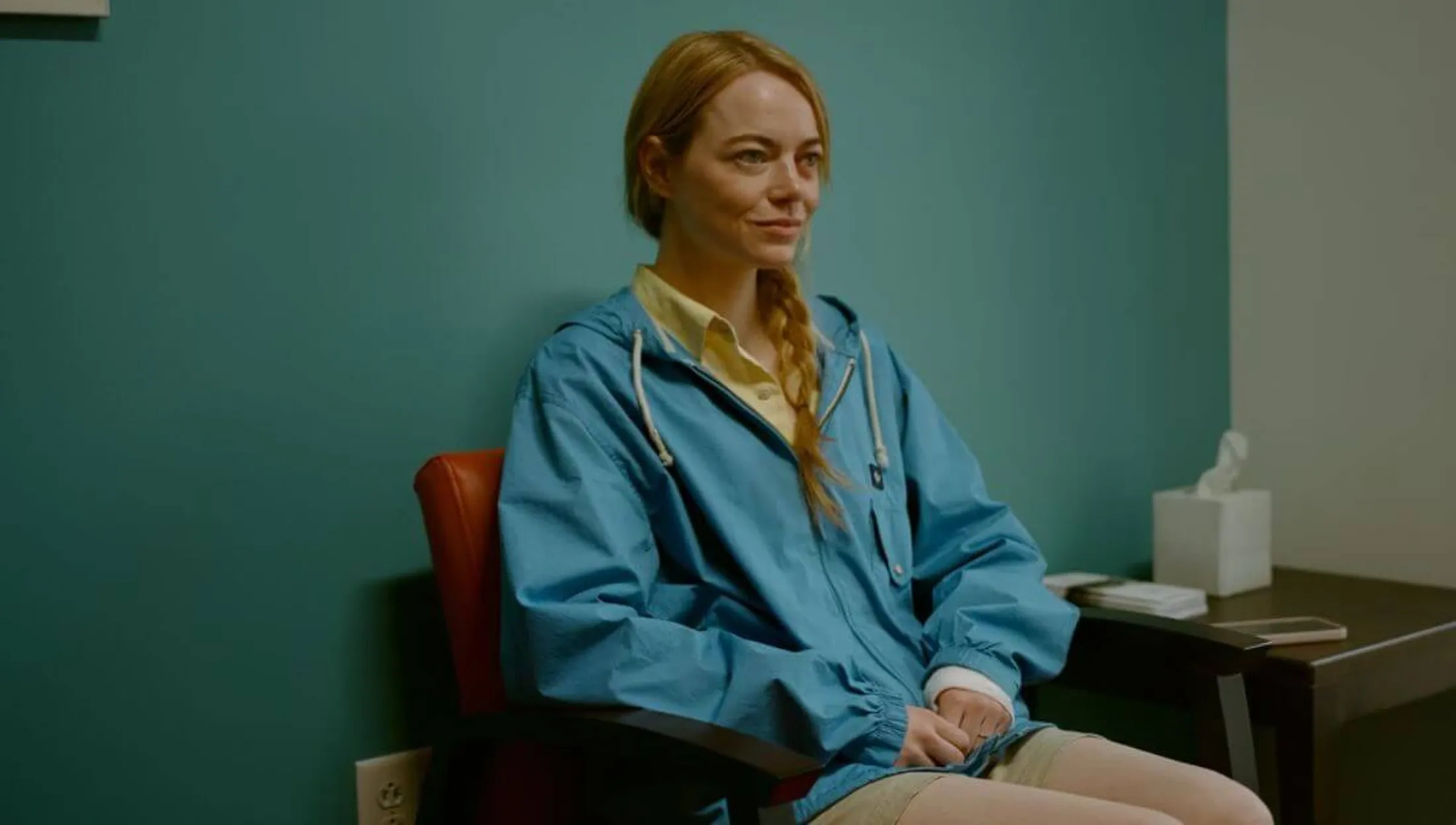 Emma Stone em Tipos de Gentileza sentada com jaqueta azul e sorridente.