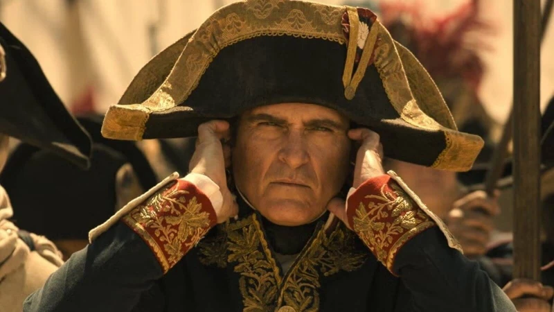 Joaquin Phoenix como Napoleão em filme de 2023. Ator aparece com chapéu, roupas de época e os dedos tapando os ouvidos.