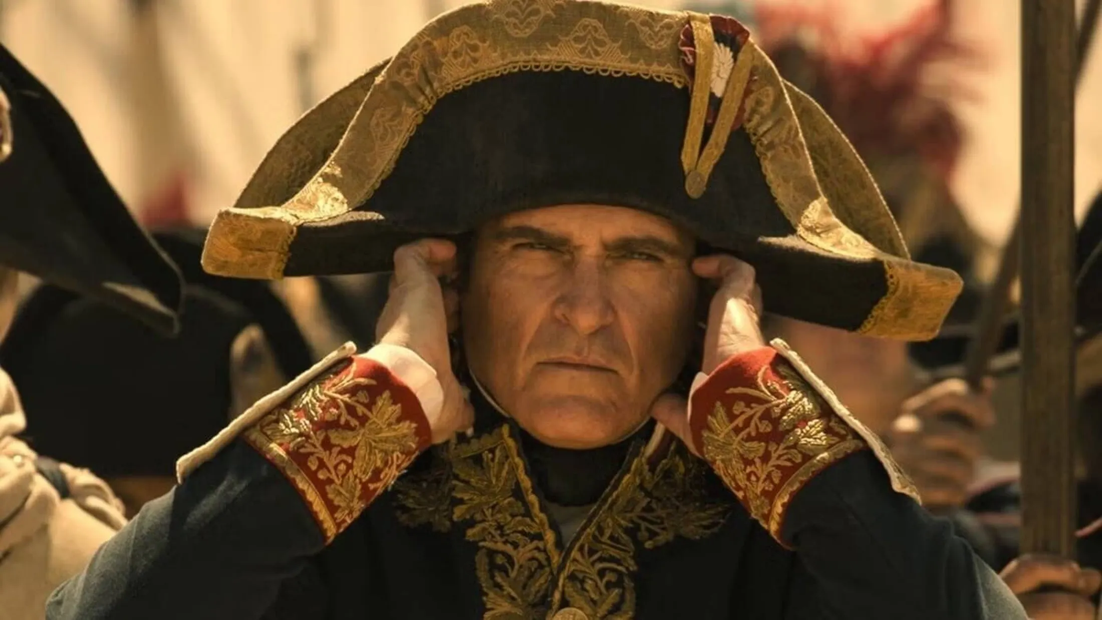 Joaquin Phoenix como Napoleão em filme de 2023. Ator aparece com chapéu, roupas de época e os dedos tapando os ouvidos.